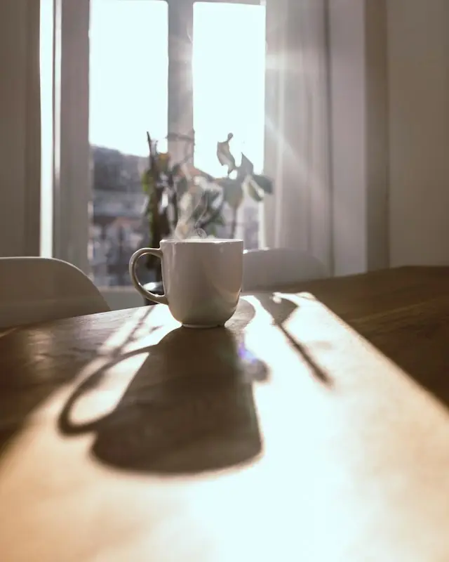 A steaming white mug on a wooden table in front of a sunlit window